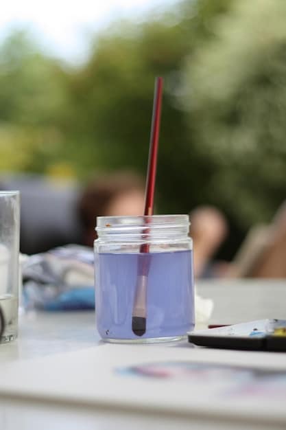 A close-up shot of a small, open watercolor pan set with a water brush next to it, on a wooden table, with a blurred background of a scenic landscape.