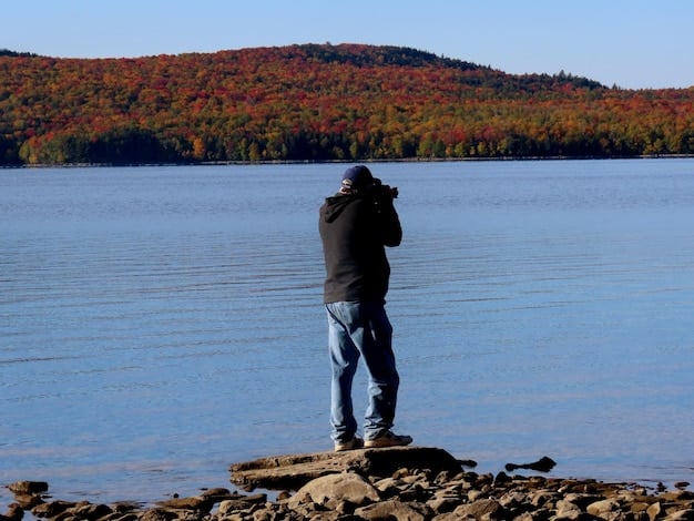A photographer standing by a large body of water, possibly a lake in a national park like Lake Louise in Banff, Canada (or a similar US lake like Lake Tahoe), with mountains reflecting in the water during golden hour. The photographer has a camera on a tripod.